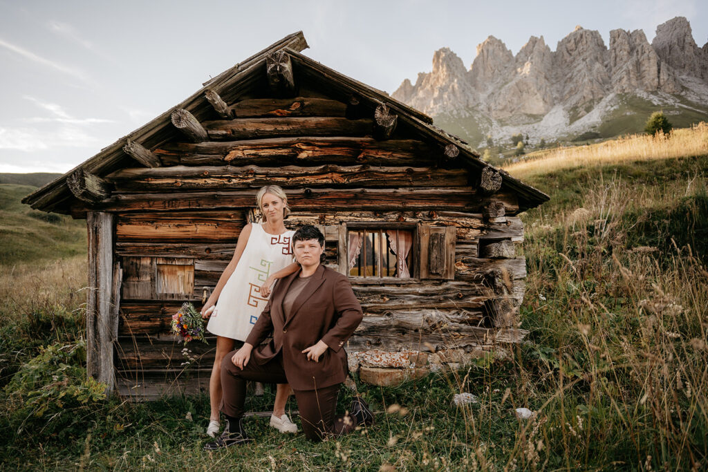 Two people pose by rustic cabin, mountains in background.