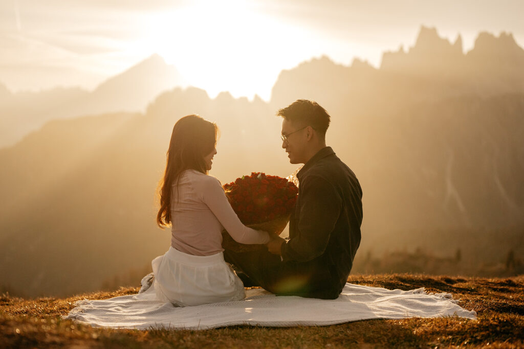 Couple sitting with roses at sunset on blanket.