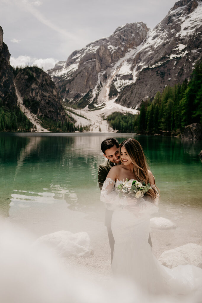 Couple embraces by mountain lake on wedding day.