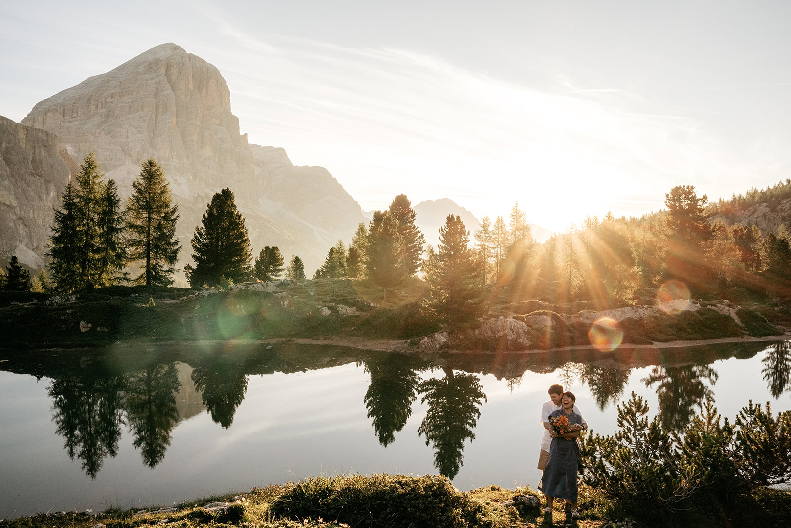 Couple embraces by mountain lake at sunrise.