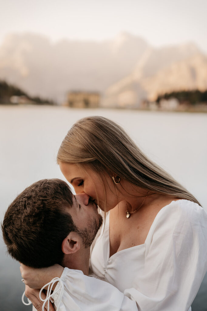 Couple kissing by a lake with mountains.