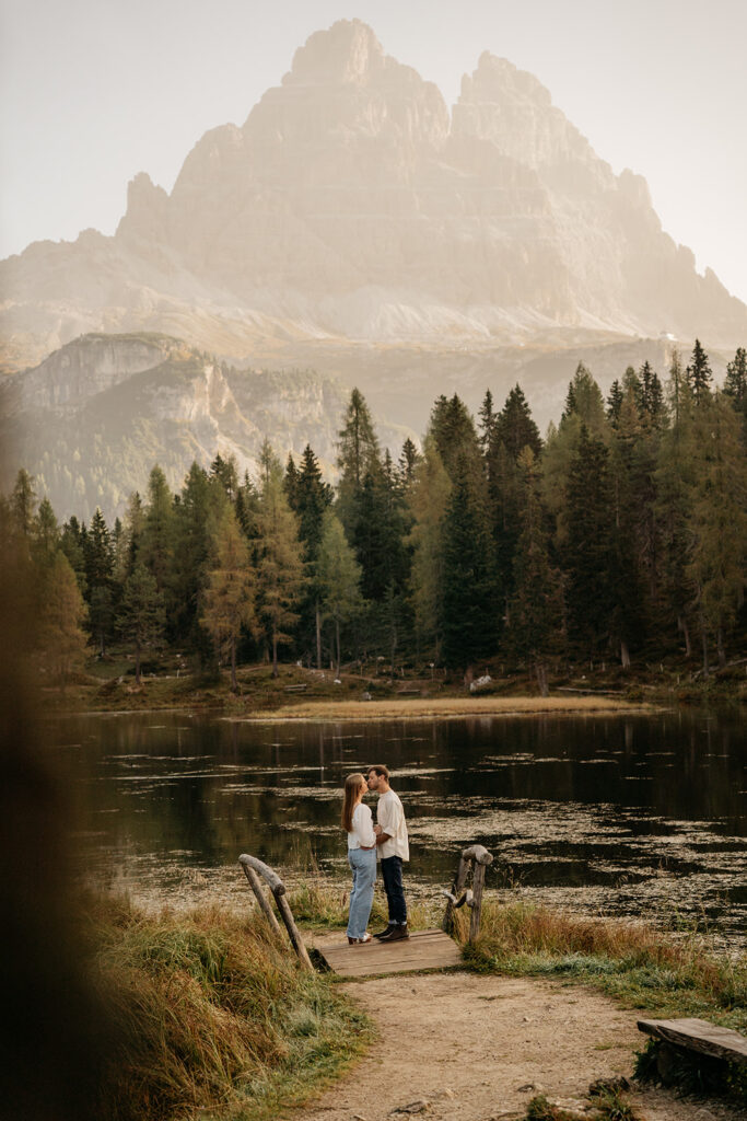 Couple embracing by lake with mountain backdrop.