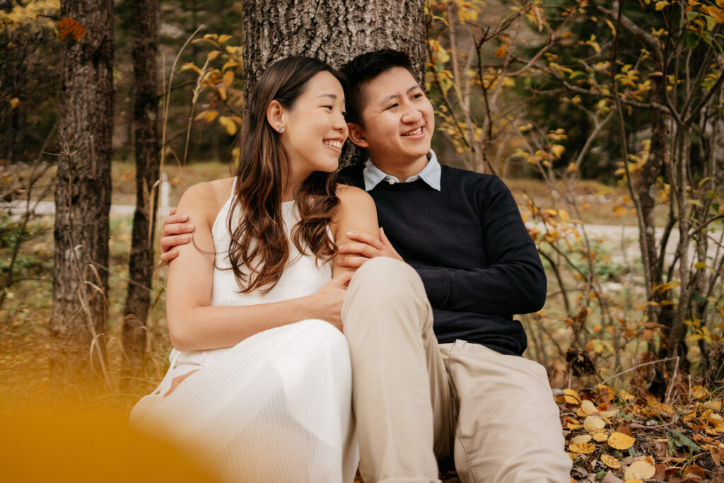 Couple smiling, sitting by a tree in autumn.