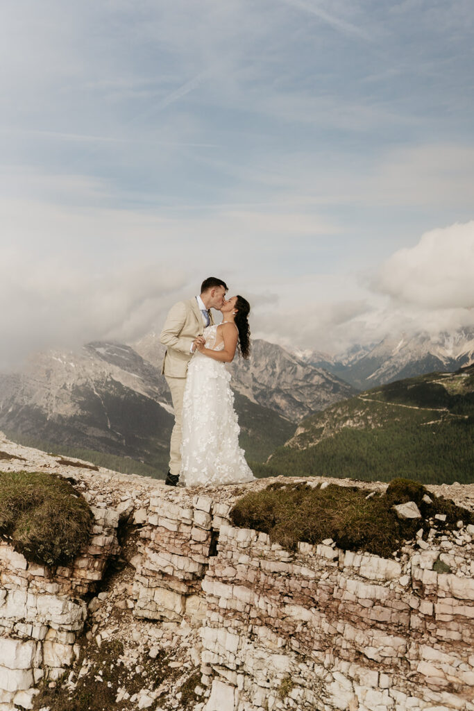 Couple kissing on mountain cliff, wedding attire.