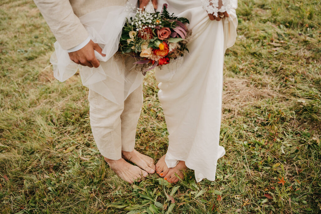 Bride and groom barefoot with wedding bouquet on grass.