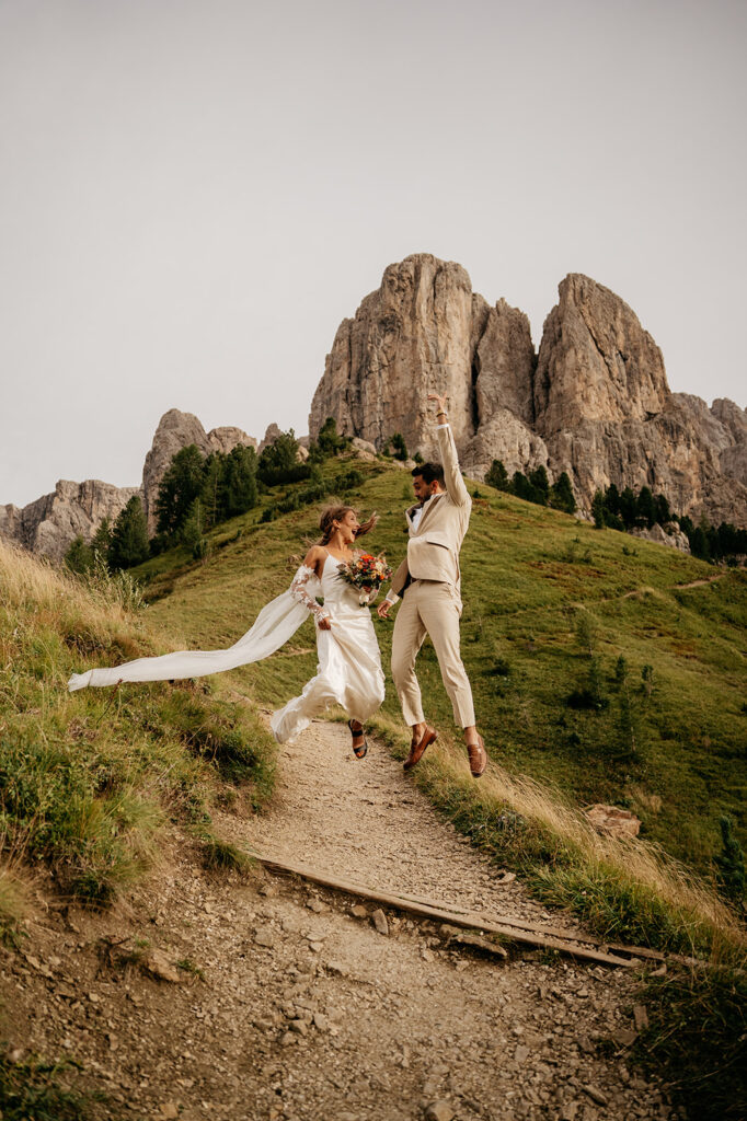 Couple jumping on mountain trail in wedding attire.