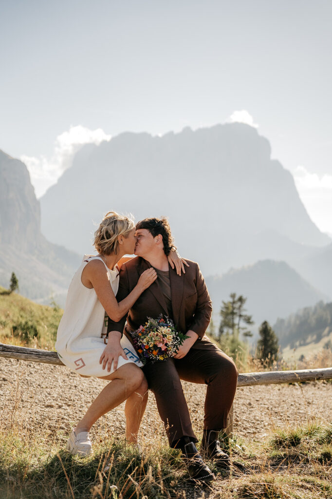 Couple kissing on mountain path, holding bouquet.