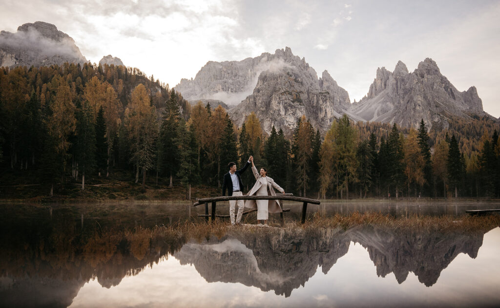 Couple on bridge with mountains and lake reflection.