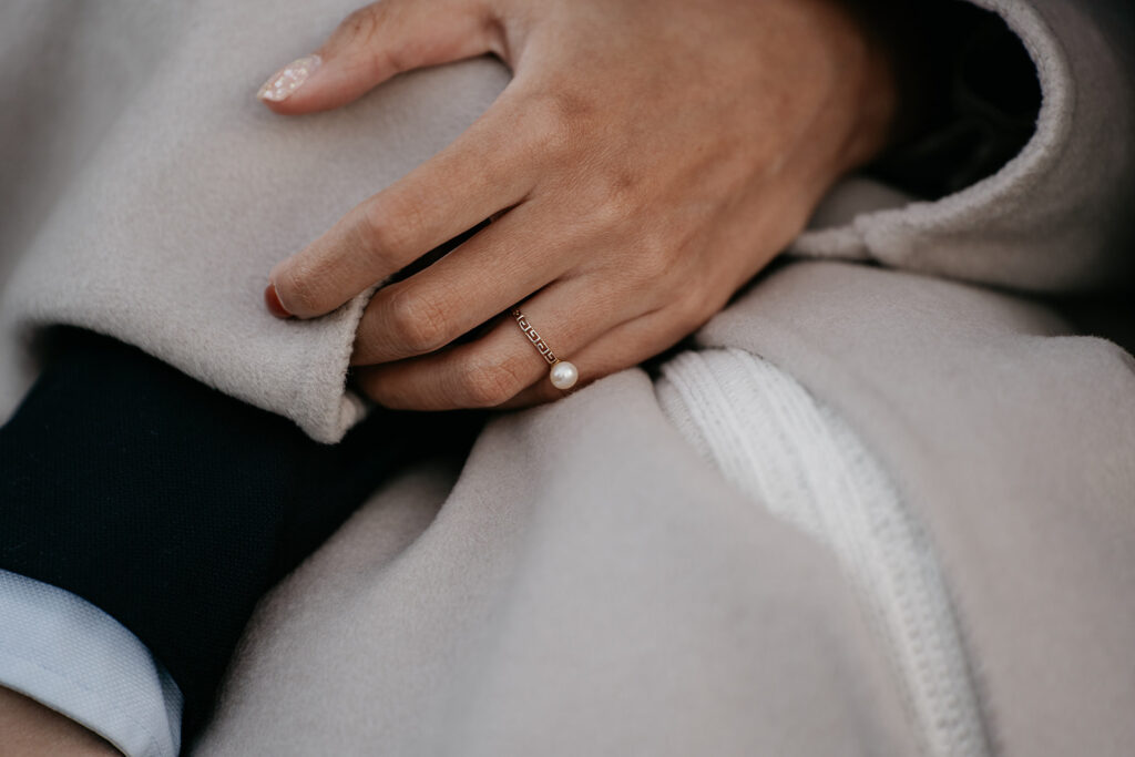 Hand wearing pearl ring on beige coat.