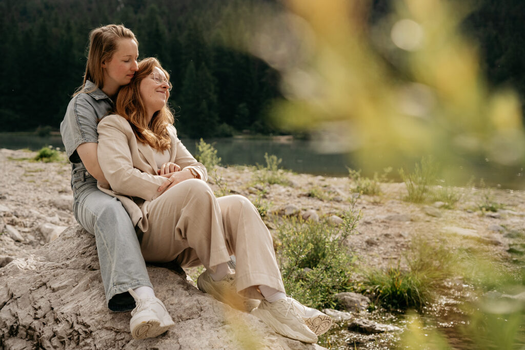 Couple embraces on rocky riverbank with forest backdrop.