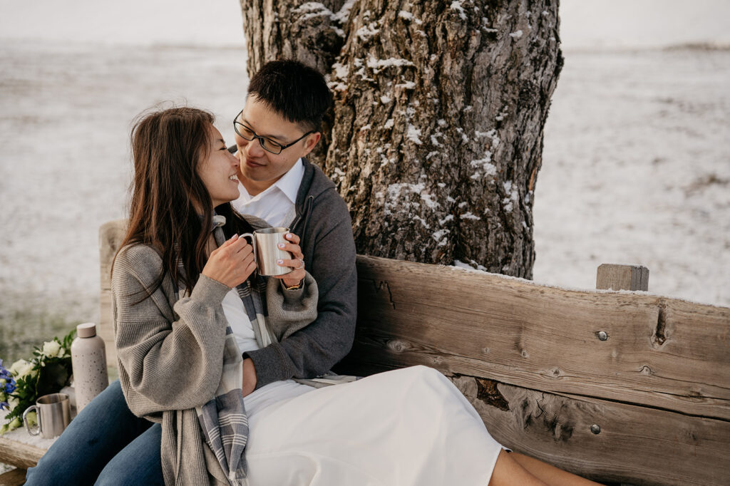 Couple enjoying hot drinks by a snowy tree.