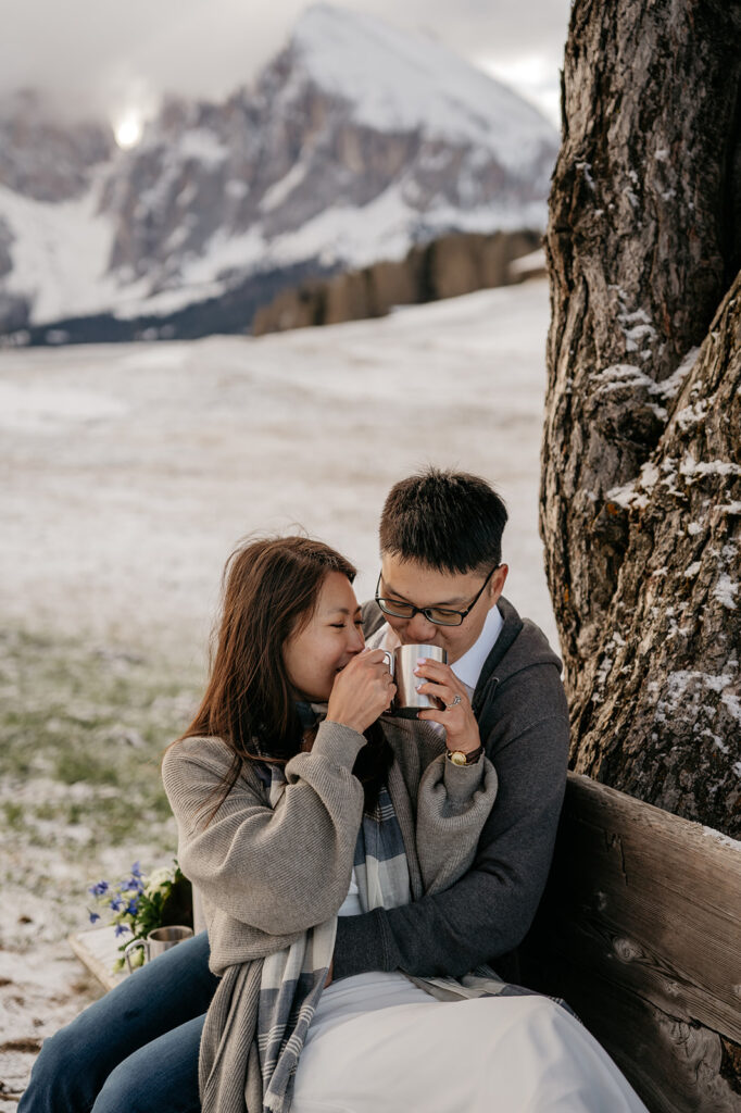 Couple enjoys hot drinks with snowy mountain backdrop.