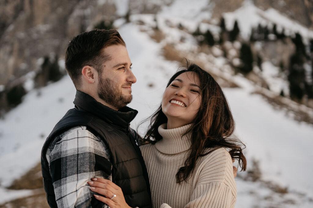 Smiling couple embraces in snowy mountain landscape.