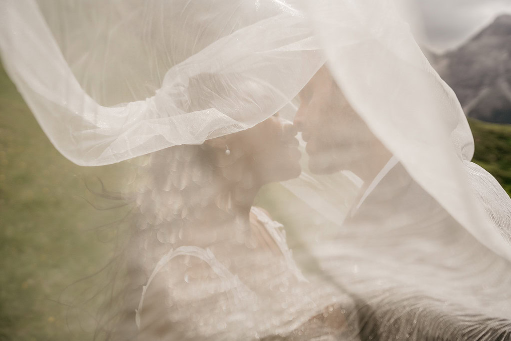 Couple kissing under wedding veil outdoors.