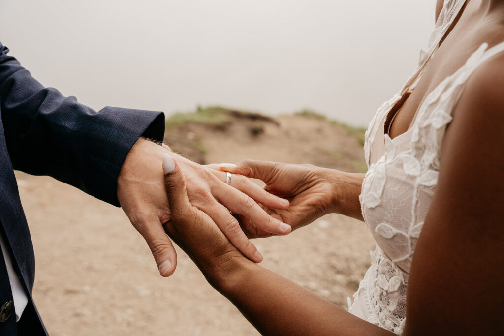 Bride placing ring on groom's finger outdoors.