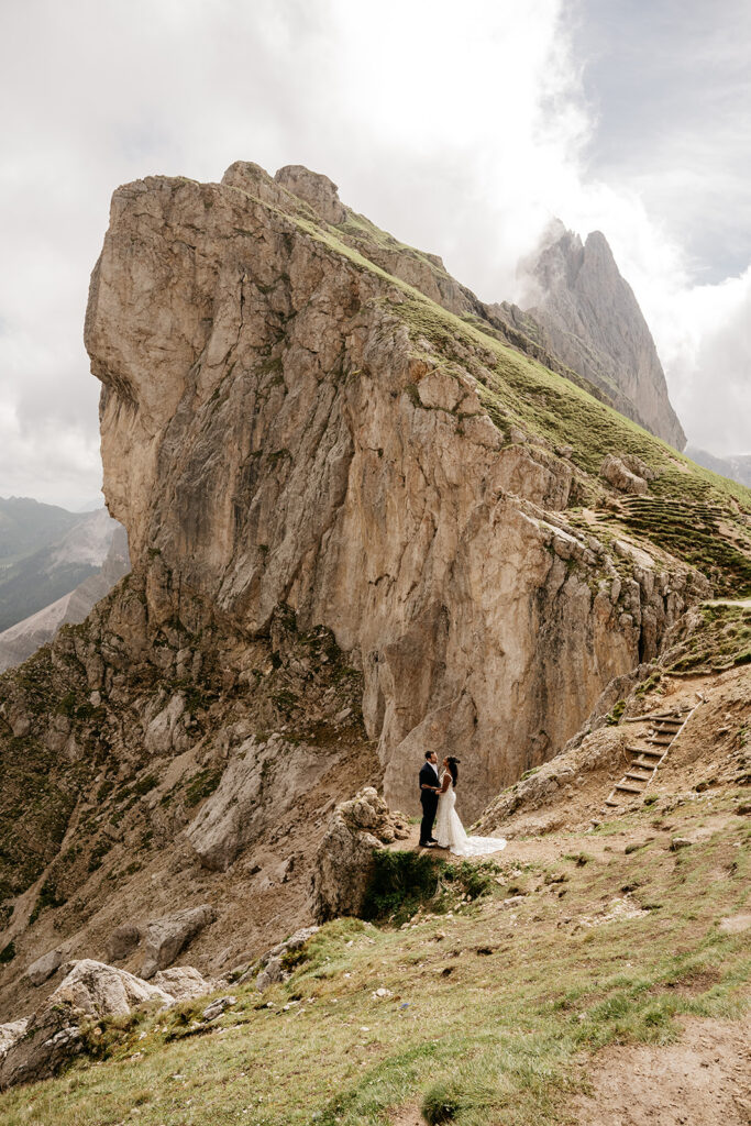 Couple stands on mountain ledge with rocky backdrop.