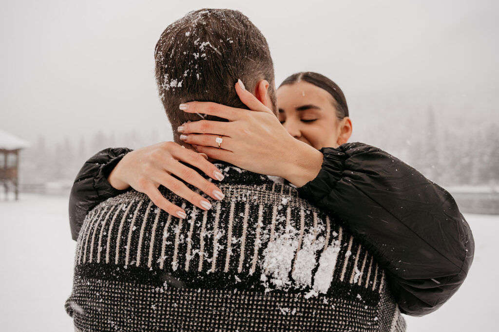 Couple embracing in snowy landscape, showing engagement ring.