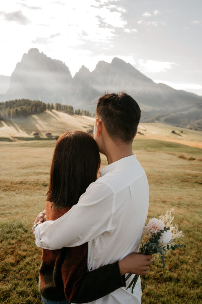 Couple embraces with mountain view, holding flowers.