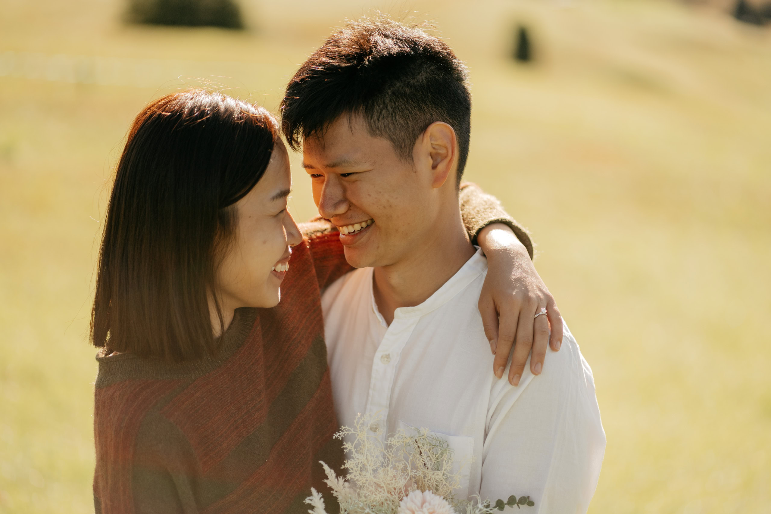 Smiling couple embracing outdoors, holding flowers.