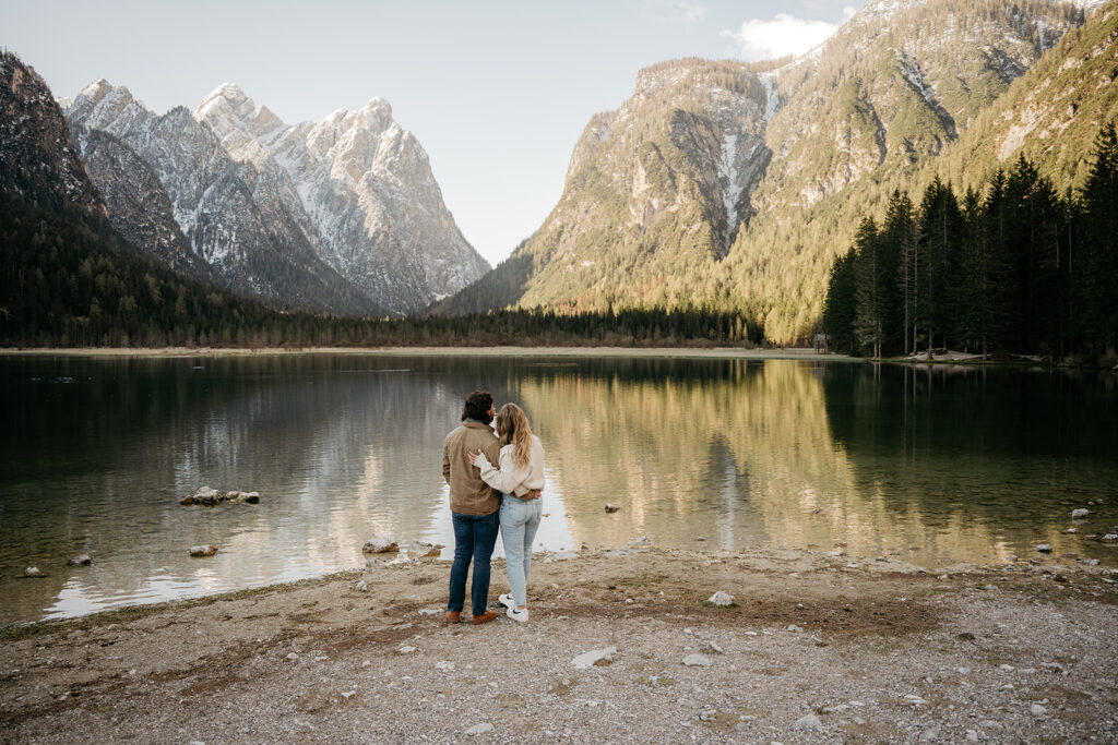 Couple embraces by lake with mountain backdrop.