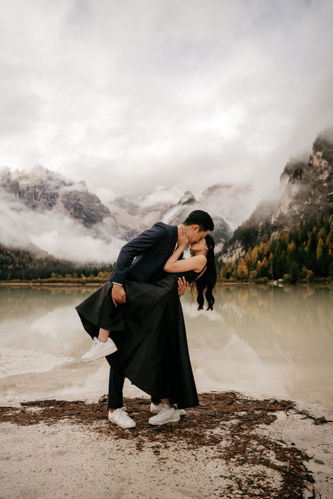 Couple kissing by mountain lake under cloudy sky.