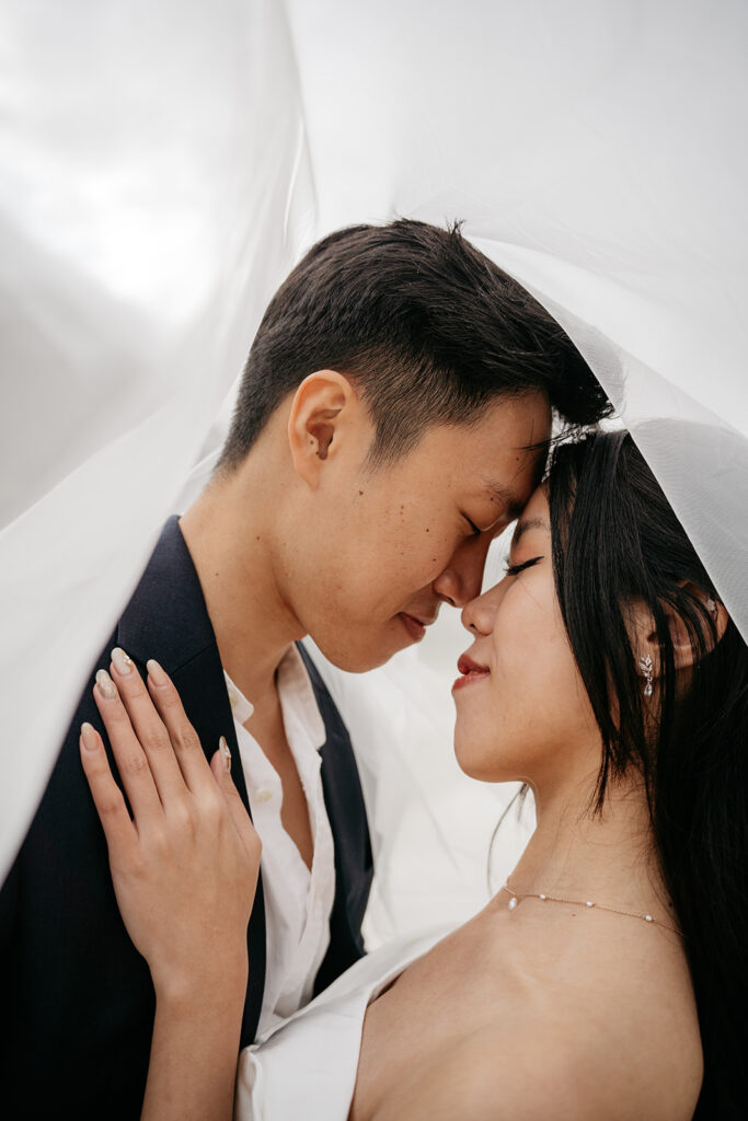 Couple embracing under white veil, eyes closed.