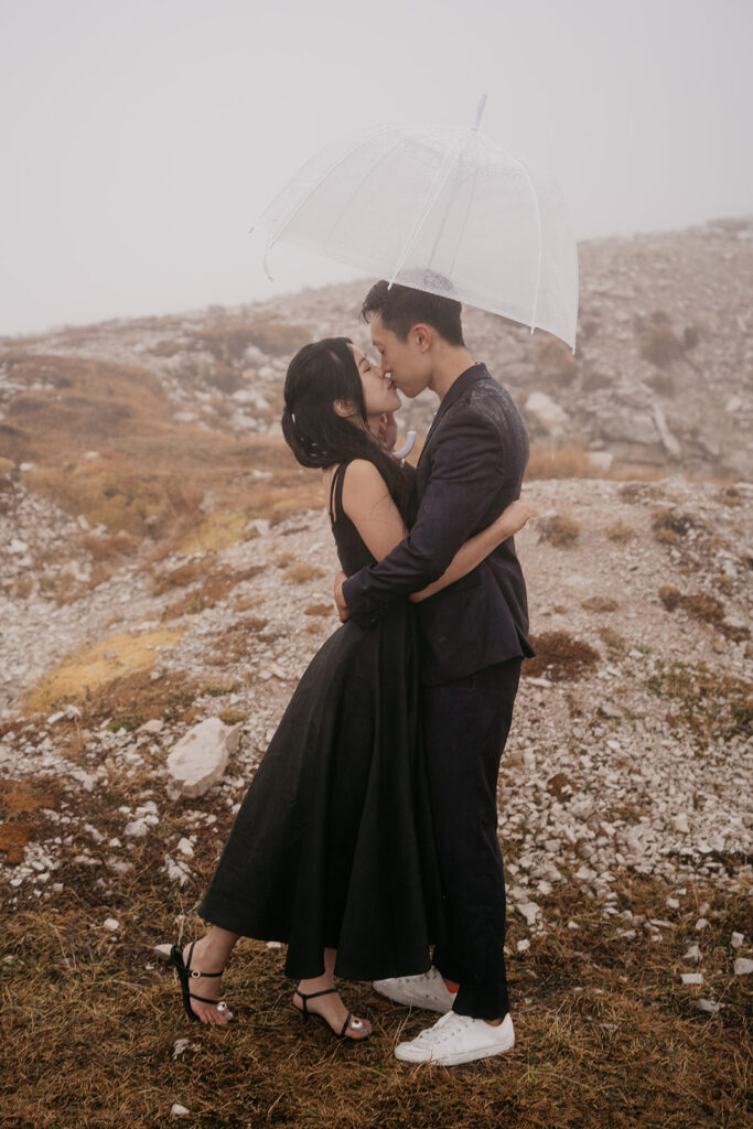 Couple kissing under umbrella in misty landscape.