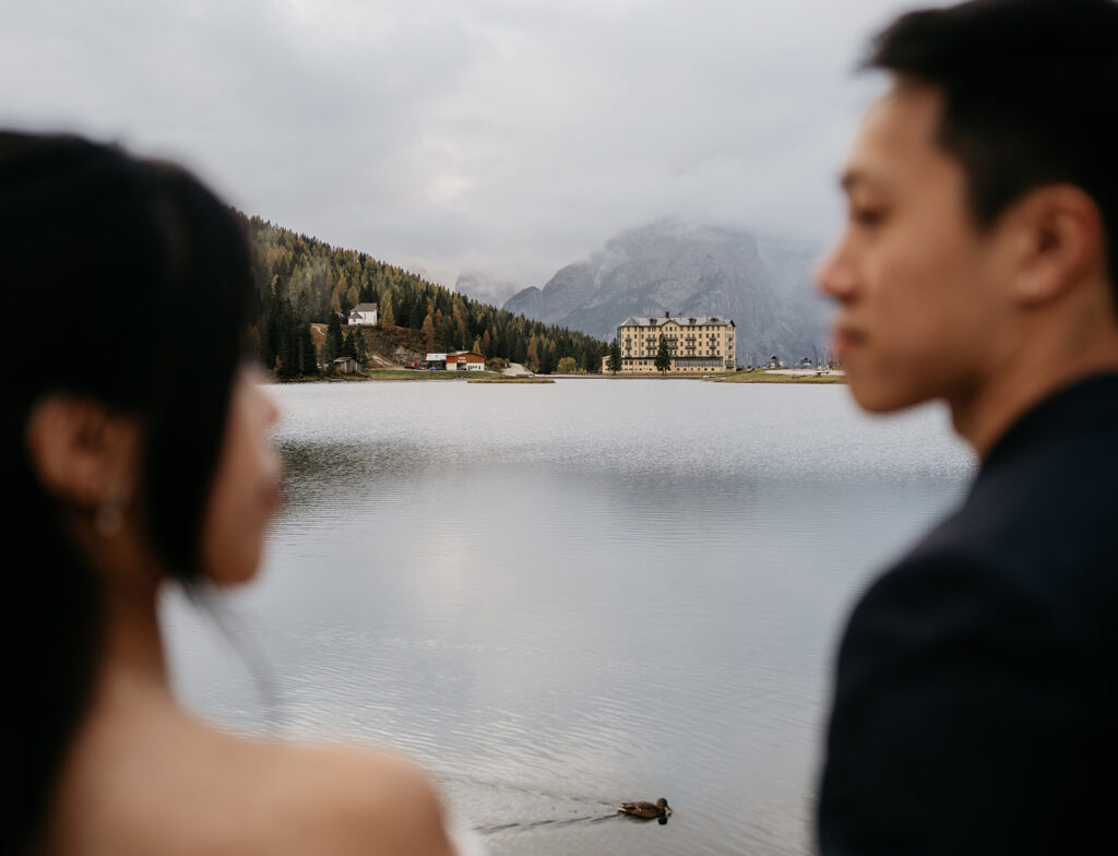 Couple by serene lake with distant mountain hotel.