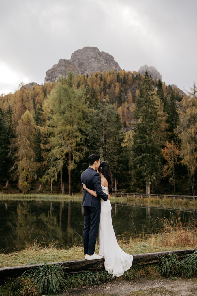 Couple embracing by forested lake and mountain backdrop.