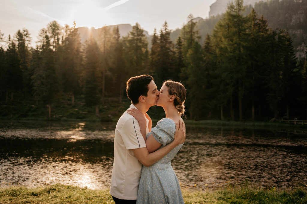 Couple kissing by a forested lake at sunset.