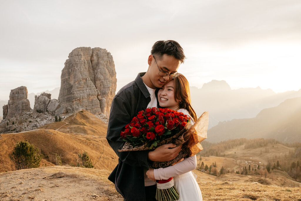 Couple embraces with roses in mountainous landscape.