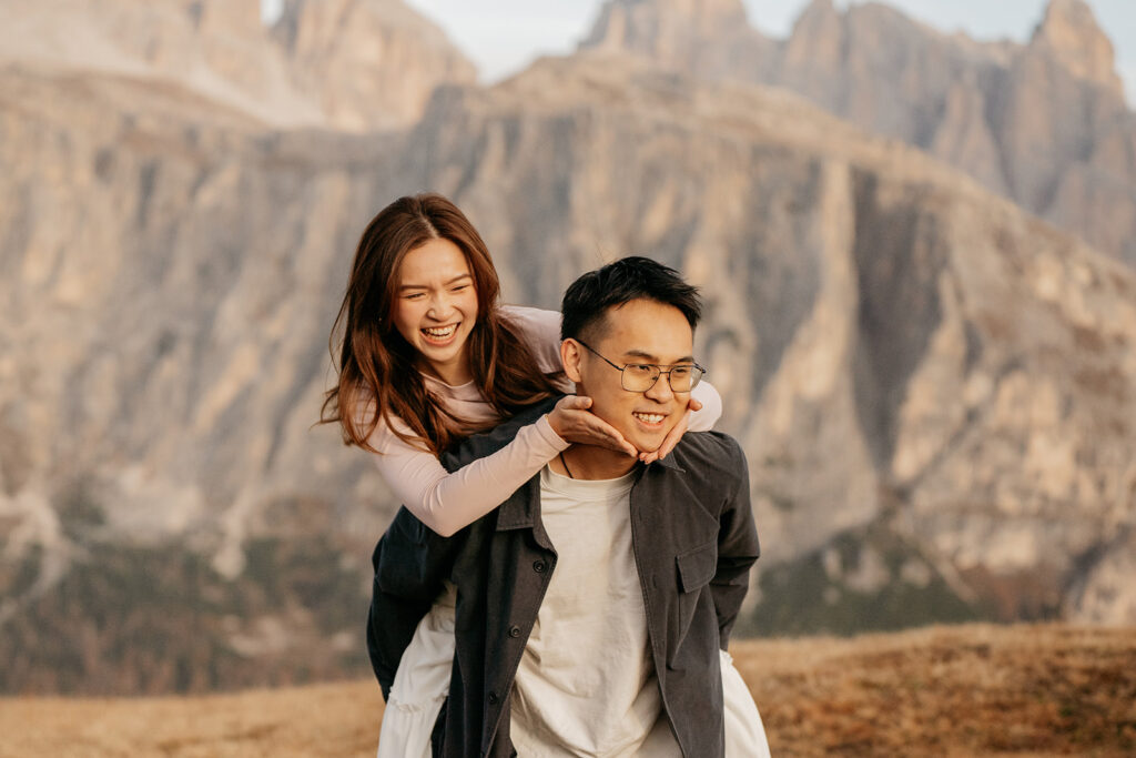 Couple piggybacking with mountain backdrop, smiling joyfully.