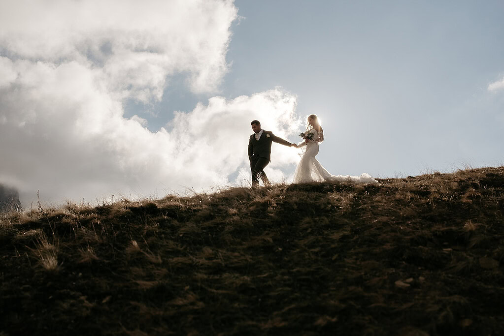 Bride and groom walking on hill under clouds.