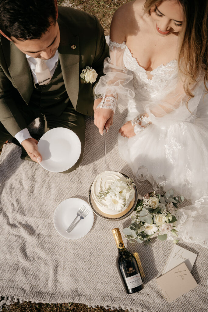 Couple cutting wedding cake outdoors with champagne.