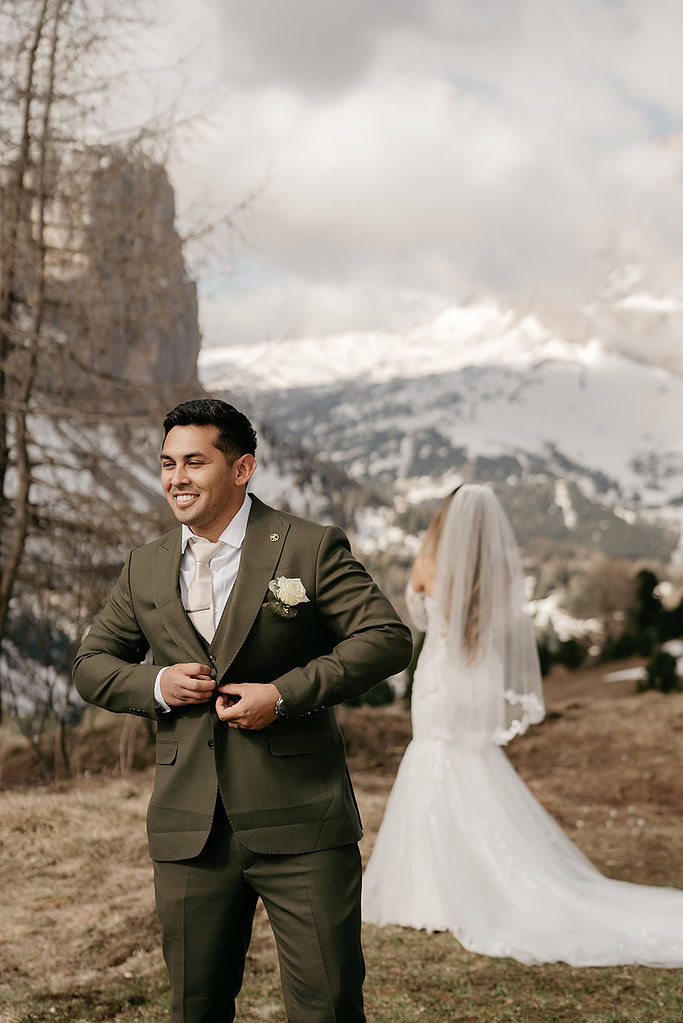 Groom smiling, bride in background, mountain landscape.