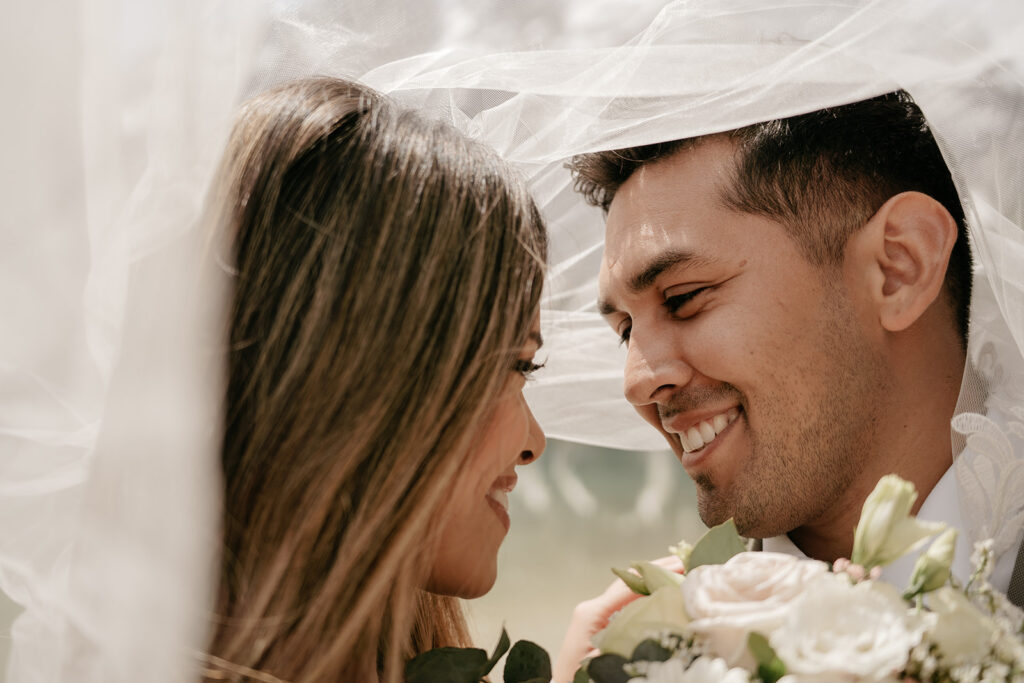 Smiling couple under wedding veil with flowers.