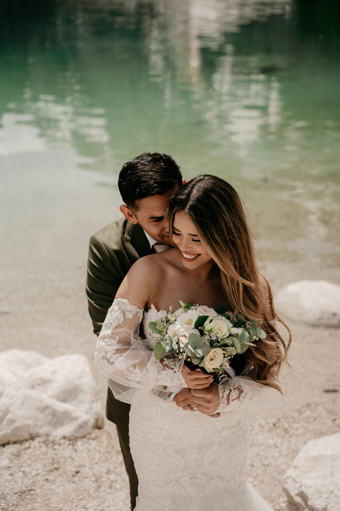 Bride with bouquet embraced by groom at lake.