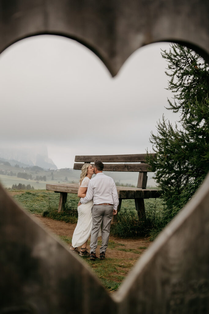 Couple kissing by wooden bench, framed by heart shape.