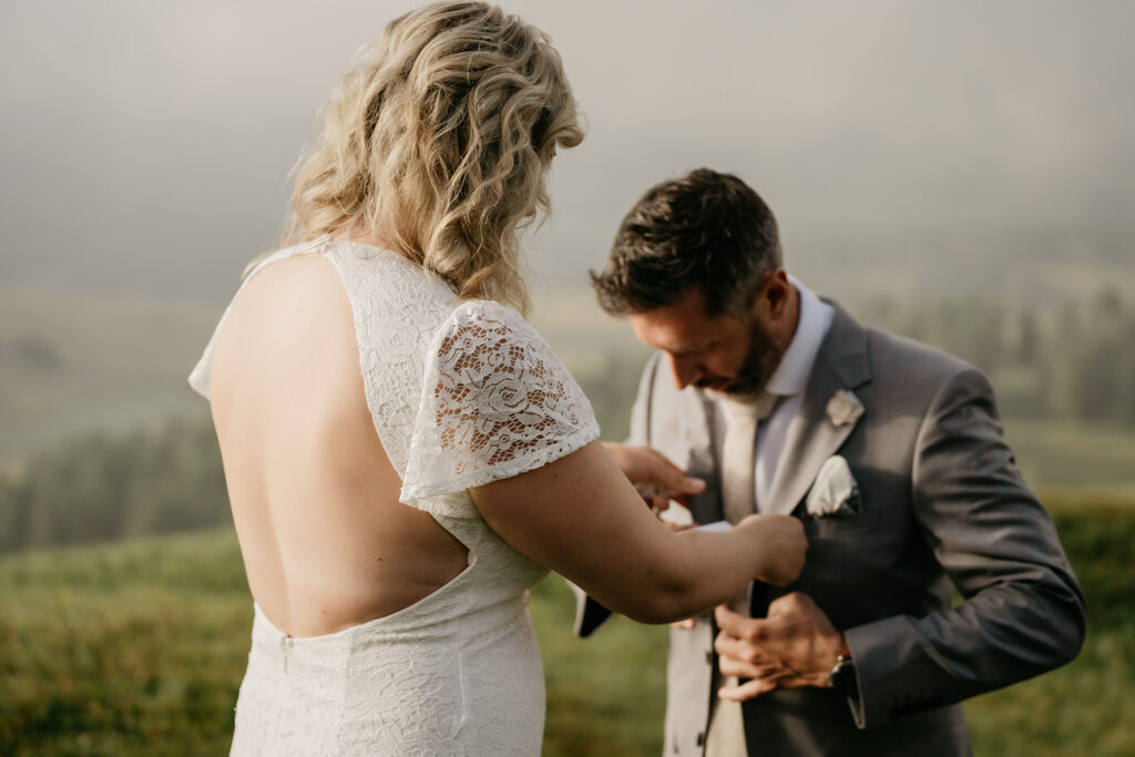 Bride helps groom with suit in nature setting.