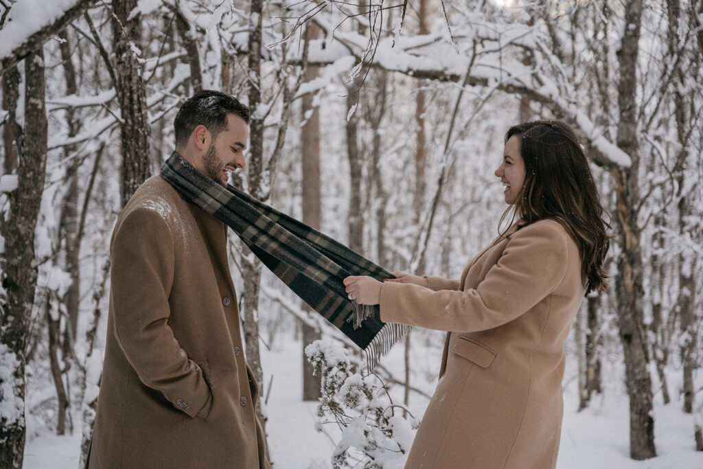 Couple in snowy forest sharing scarf joyfully.