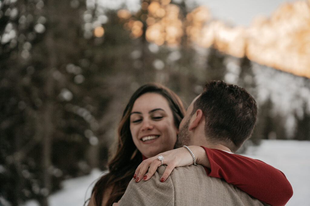 Couple embracing in snowy forest with engagement ring.