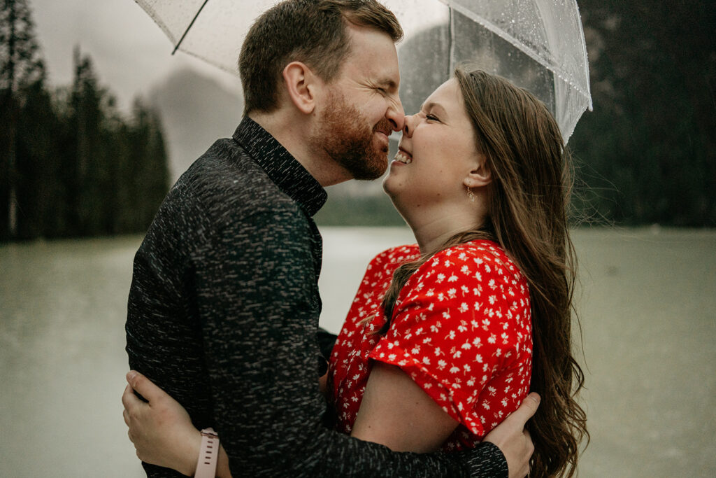 Couple smiling under clear umbrella by a lake.