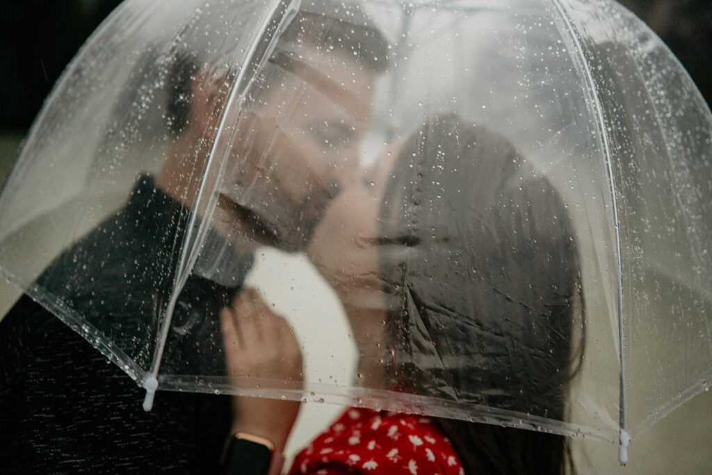 Couple kissing under clear umbrella in rain.