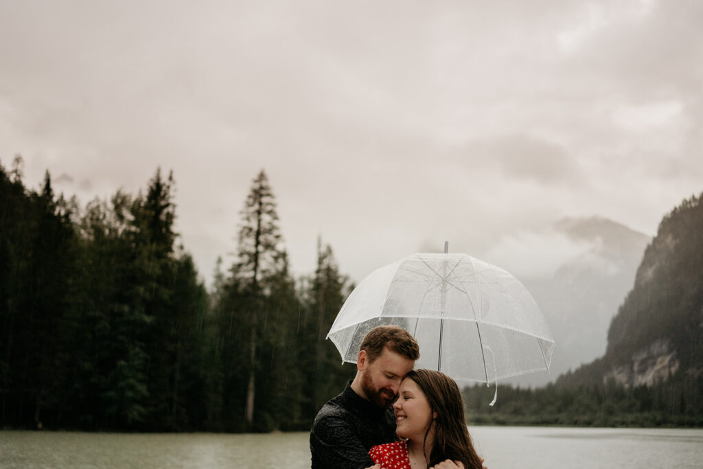 Couple hugging under umbrella by a lake.