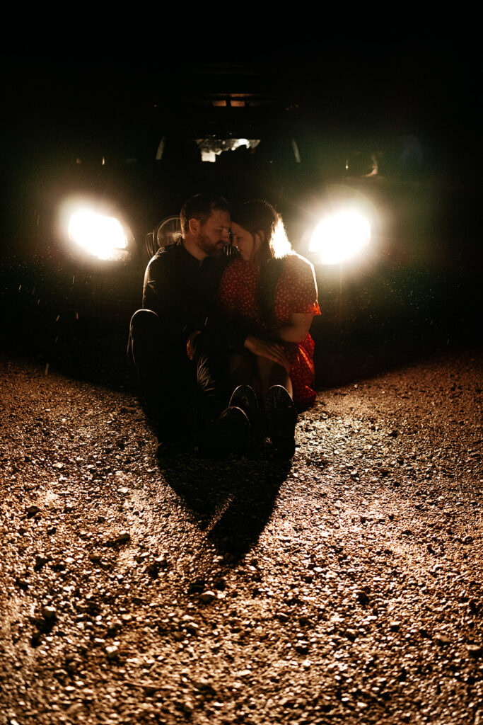 Couple sitting by car headlights at night