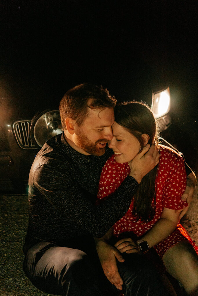 Couple embracing at night near car headlights.