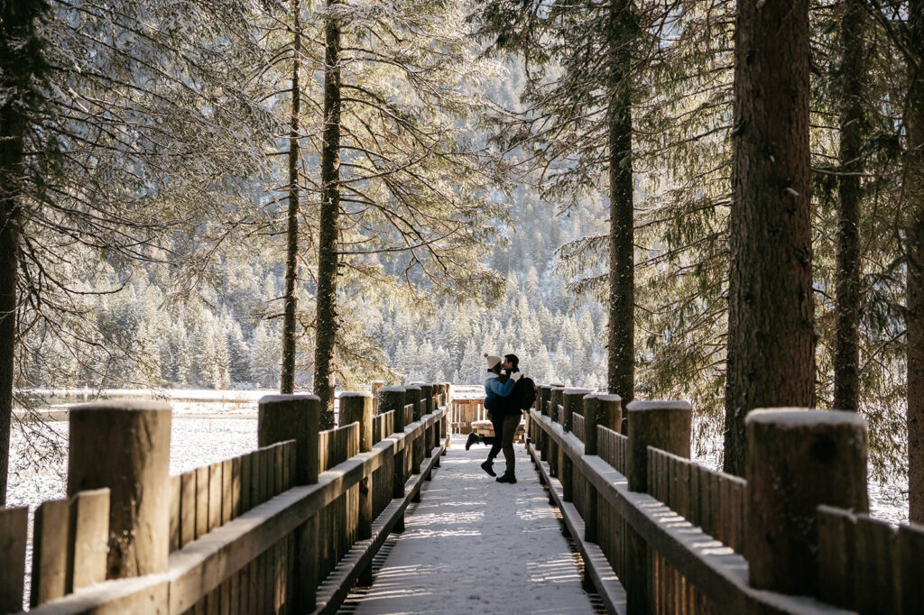 Couple embraces on snowy wooded bridge.