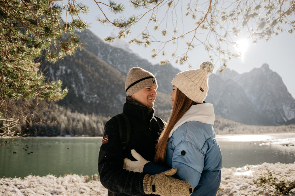 Couple in winter coats near snowy mountain lake.