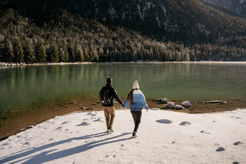 Couple holding hands by snowy lake, forest background.