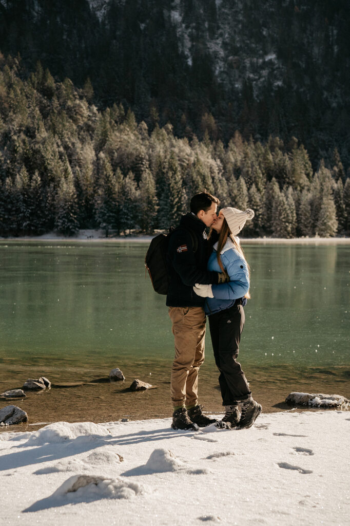 Couple kissing by a snowy lake with trees.